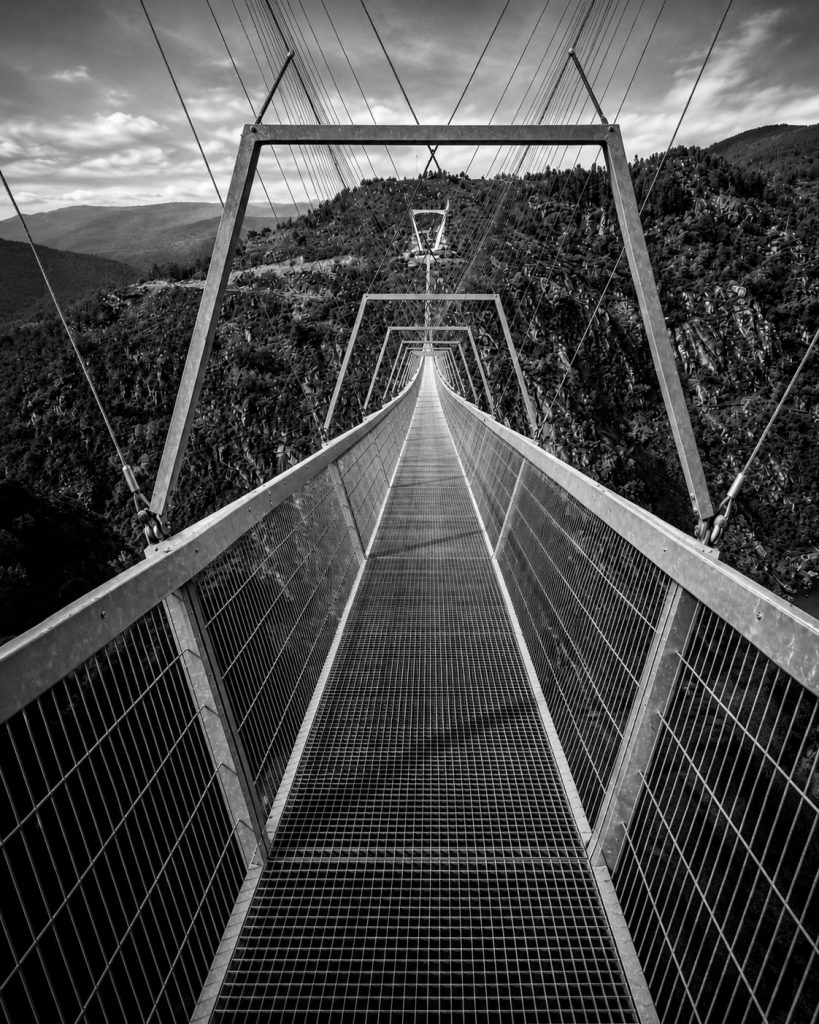 Imagem vertical em preto e branco da Ponte 516 Arouca vista a partir do tabuleiro vazio, com passadiço metálico em grelha, guardas laterais, cabos de suspensão e estruturas repetidas a criar uma forte perspectiva central sobre uma paisagem montanhosa.