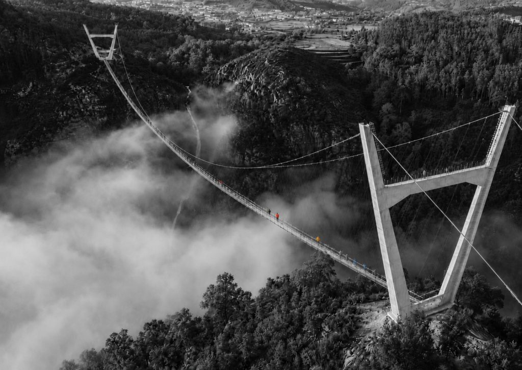 Imagem aérea quase monocromática da Ponte 516 Arouca a atravessar um vale coberto de neblina, com a paisagem e a estrutura em preto e branco e poucos visitantes destacados a cores sobre o tabuleiro da ponte.