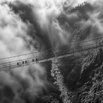 Imagem a preto e branco da Ponte 516 Arouca a atravessar o vale do rio Paiva entre neblina densa e encostas rochosas, com vários visitantes agrupados numa secção da ponte suspensa.