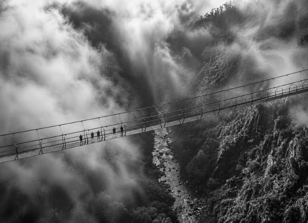 Imagem a preto e branco da Ponte 516 Arouca a atravessar o vale do rio Paiva entre neblina densa e encostas rochosas, com vários visitantes agrupados numa secção da ponte suspensa.