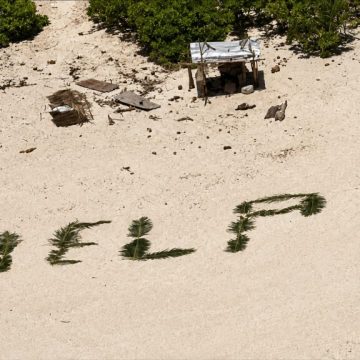 Vista aérea de uma praia remota com vegetação tropical, onde a palavra “HELP” aparece escrita na areia com folhas de palmeira, simbolizando o pedido de socorro que ajudou três homens a serem resgatados numa ilha do Pacífico.