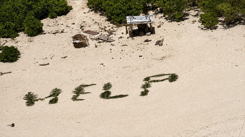 Vista aérea de uma praia remota com vegetação tropical, onde a palavra “HELP” aparece escrita na areia com folhas de palmeira, simbolizando o pedido de socorro que ajudou três homens a serem resgatados numa ilha do Pacífico.