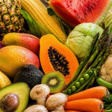 Composição colorida e aproximada de frutas e legumes frescos da lista Quinze Limpos, incluindo ananás, milho, melancia, abacate, papaia, couve-flor, couve-repolho, cebolas, espargos, cenouras, cogumelos, kiwi e bananas, com gotas de água sobre os alimentos.