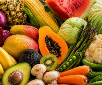 Composição colorida e aproximada de frutas e legumes frescos da lista Quinze Limpos, incluindo ananás, milho, melancia, abacate, papaia, couve-flor, couve-repolho, cebolas, espargos, cenouras, cogumelos, kiwi e bananas, com gotas de água sobre os alimentos.