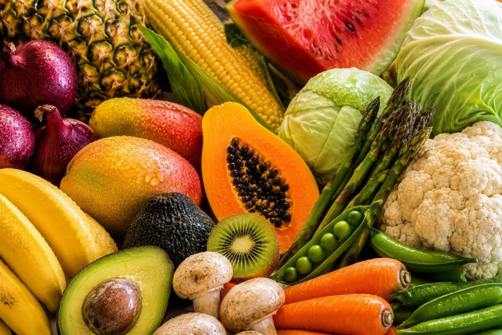 Composição colorida e aproximada de frutas e legumes frescos da lista Quinze Limpos, incluindo ananás, milho, melancia, abacate, papaia, couve-flor, couve-repolho, cebolas, espargos, cenouras, cogumelos, kiwi e bananas, com gotas de água sobre os alimentos.