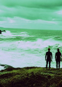 Dois surfistas em fato de neoprene observam o mar revolto a partir de uma zona segura na costa.
