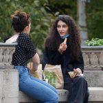 Duas jovens conversam e riem enquanto almoçam juntas num banco de jardim