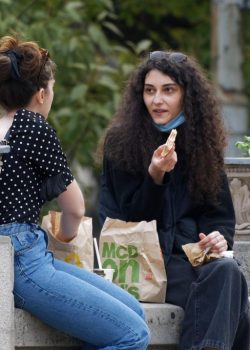 Duas jovens conversam e riem enquanto almoçam juntas num banco de jardim