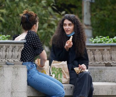 Duas jovens conversam e riem enquanto almoçam juntas num banco de jardim