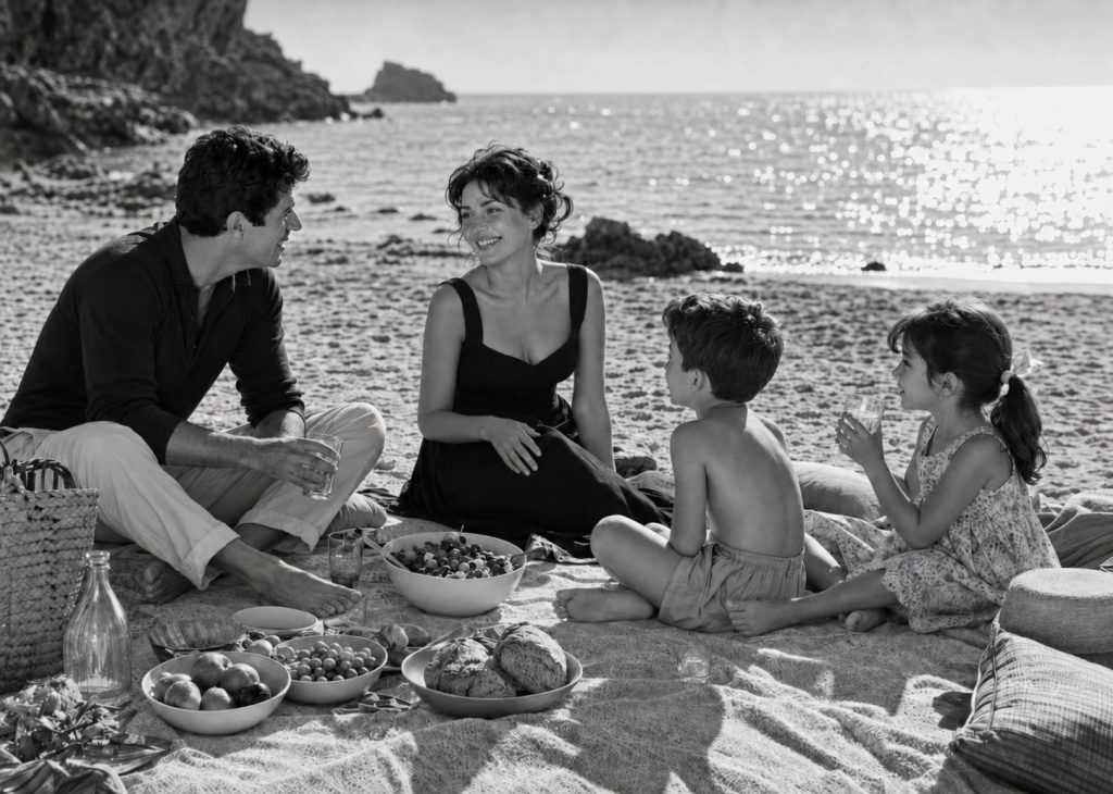 Família sentada numa manta na praia a partilhar um almoço com saladas, fruta e pão, numa imagem a preto e branco com atmosfera de cinema italiano clássico.
