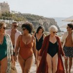 Grupo diverso de mulheres e homens de várias idades e corpos a caminhar junto ao mar, com fatos de banho elegantes, num ambiente mediterrânico de Verão.