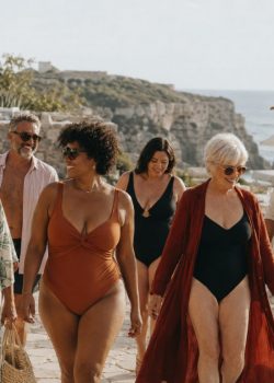Grupo diverso de mulheres e homens de várias idades e corpos a caminhar junto ao mar, com fatos de banho elegantes, num ambiente mediterrânico de Verão.