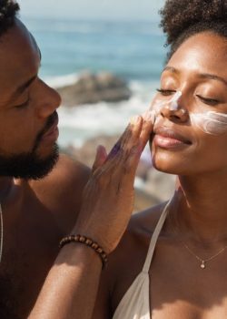 Casal negro na praia em plano aproximado, com o homem a aplicar protetor solar no rosto da mulher, enquanto ela mantém os olhos fechados, com o mar e as rochas ao fundo.