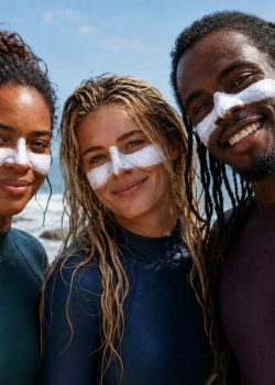 Três surfistas junto ao mar — uma mulher negra, uma mulher branca loira e um homem negro — usam fatos de surf coloridos e têm protetor solar mineral branco, espesso, espalhado nas maçãs do rosto e no nariz.