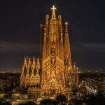 Vista noturna horizontal da Sagrada Família, em Barcelona, iluminada com destaque para a Torre de Jesus Cristo e a cruz brilhante no topo, com a cidade ao fundo.