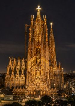 Vista noturna horizontal da Sagrada Família, em Barcelona, iluminada com destaque para a Torre de Jesus Cristo e a cruz brilhante no topo, com a cidade ao fundo.