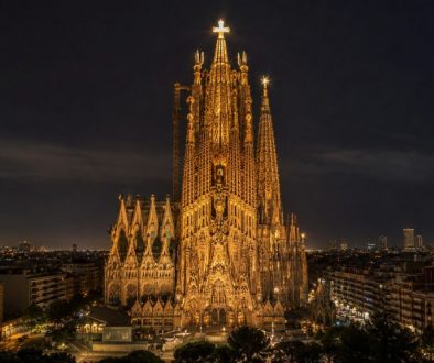 Vista noturna horizontal da Sagrada Família, em Barcelona, iluminada com destaque para a Torre de Jesus Cristo e a cruz brilhante no topo, com a cidade ao fundo.