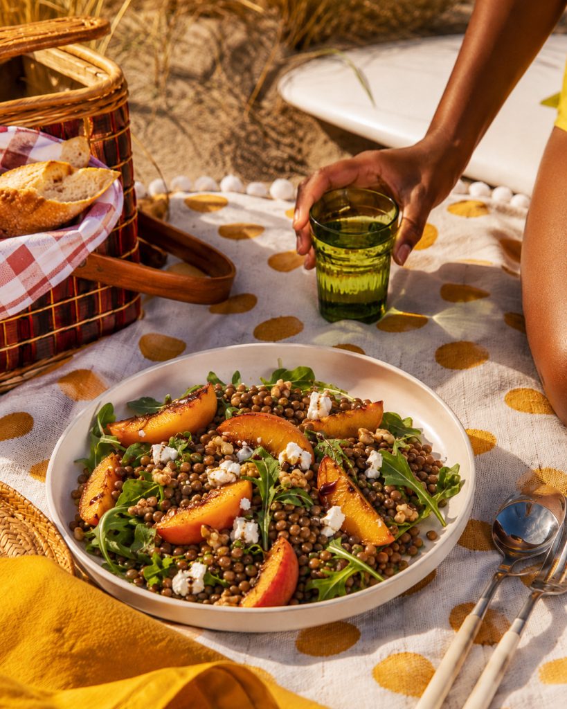 Salada vegetariana de lentilhas com pêssego assado, rúcula, nozes e queijo de cabra numa taça clara sobre uma manta de praia amarela, com copo verde, talheres e cesto de piquenique ao lado.