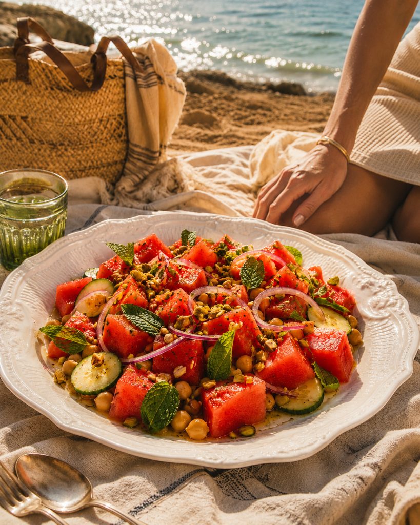 Prato branco com salada de melancia, grão-de-bico, pepino, cebola roxa, hortelã e pistácios sobre uma manta na praia, com copo verde, talheres e o mar ao fundo.