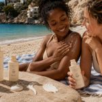 Duas mulheres jovens na praia, uma negra e uma branca, relaxam sobre uma toalha junto ao mar, com protetor solar aplicado no ombro; em primeiro plano, vários frascos de protetor solar mostram diferentes níveis de FPS, incluindo 15, 30, 50 e 50+.