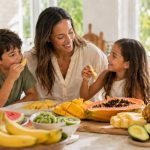Mãe com dois filhos sentados à ilha da cozinha, a comer frutas frescas da lista dos Quinze Limpos, incluindo ananás, banana, papaia, melancia, manga, kiwi e abacate, numa cozinha luminosa e familiar.