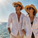 Homem e mulher junto ao mar, ambos com chapéus de palha e óculos de sol, usando camisas leves brancas sobre roupa de praia, numa paisagem costeira luminosa com o sol alto e a água a brilhar ao fundo.