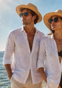 Homem e mulher junto ao mar, ambos com chapéus de palha e óculos de sol, usando camisas leves brancas sobre roupa de praia, numa paisagem costeira luminosa com o sol alto e a água a brilhar ao fundo.